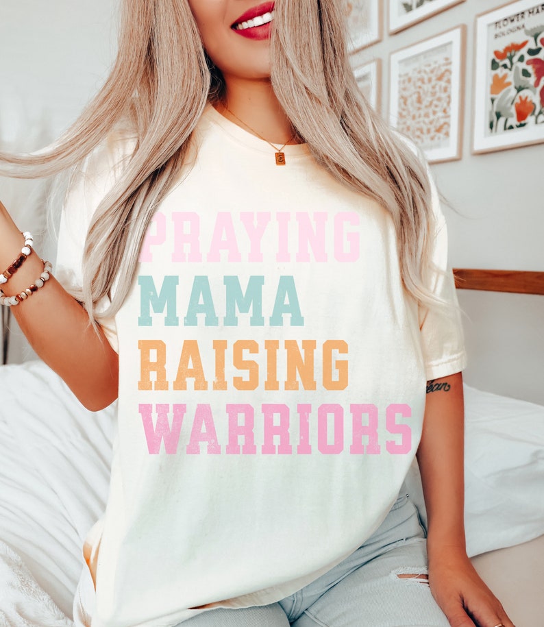 May include: A white t-shirt with the text 'Praying Mama Raising Warriors' in pink, blue, and orange lettering. Praying Mama, Comfort Colors Tshirt, Christian Shirts, Mama Shirt, Mom Shirt, Mothers Day Gift, Gifts For Mom, Christian Apparel, Gift For Wife, Religious Shirt, Religious Gifts for Her, New Mom Gift. Mom Gifts, Christian Gifts