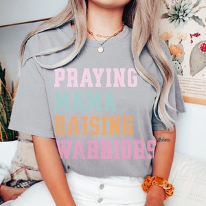 May include: A gray t-shirt with the text 'Praying Mama Raising Warriors' in pink, blue, and orange lettering. Praying Mama, Comfort Colors Tshirt, Christian Shirts, Mama Shirt, Mom Shirt, Mothers Day Gift, Gifts For Mom, Christian Apparel, Gift For Wife, Religious Shirt, Religious Gifts for Her, New Mom Gift. Mom Gifts, Christian Gifts