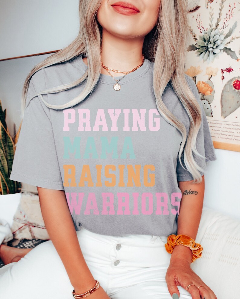 May include: A gray t-shirt with the text 'Praying Mama Raising Warriors' in pink, blue, and orange lettering. Praying Mama, Comfort Colors Tshirt, Christian Shirts, Mama Shirt, Mom Shirt, Mothers Day Gift, Gifts For Mom, Christian Apparel, Gift For Wife, Religious Shirt, Religious Gifts for Her, New Mom Gift. Mom Gifts, Christian Gifts