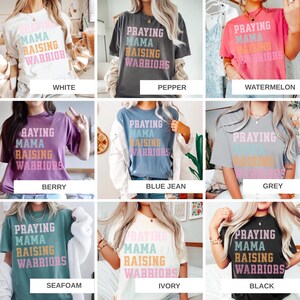 May include: A collection of t-shirts in various colors, each featuring the text "Praying Mama Raising Warriors" in a layered, colorful design. Colors include white, pepper, watermelon, berry, blue jean, grey, seafoam, ivory, and black.