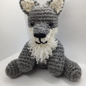 May include: A gray crocheted wolf plush toy with white fur around its face and ears. The toy has black eyes and a black nose.