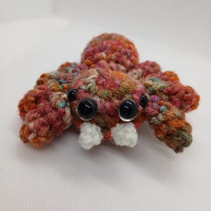 May include: A crocheted spider plush toy crafted from variegated yarn in shades of brown, orange, pink, and green. The spider features two large, glossy black plastic eyes, two smaller black side eyes, and two white crocheted fangs.