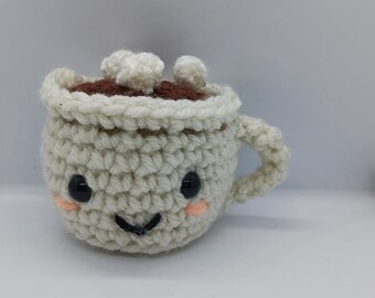 May include: A cream-colored crocheted mug with a handle, featuring a dark brown top layer representing a beverage, topped with three small white crocheted marshmallows. The mug has two black plastic eyes, a stitched black smile, and pink cheeks.
