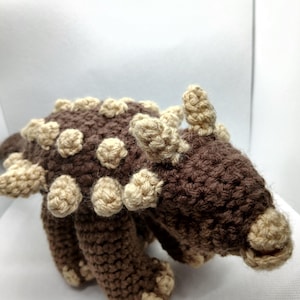 May include: A crocheted brown and tan dinosaur toy. The dinosaur has a rounded body with tan-colored bumps along its back and sides. The legs and head are also brown with tan accents. The toy is made of yarn.