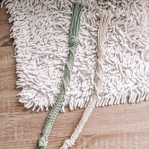 May include: Two macrame plant hangers in the image. One is a light green color, and the other is a natural beige color. Both have a knotted design and a fringe at the bottom. The hangers are lying on a white, textured rug.