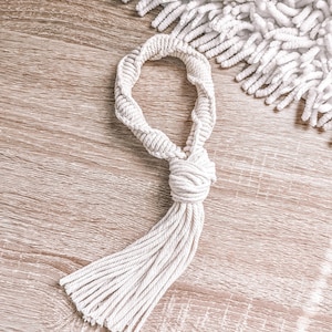 May include: Cream-colored macrame tassel with a knotted design and a looped handle. The tassel is made of multiple strands of twisted cotton rope. The background features a wooden surface and a white, fluffy textile.
