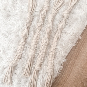 May include: Four beige macrame wall hangings with tassels. The hangings are made of natural cotton rope and have a simple, minimalist design.