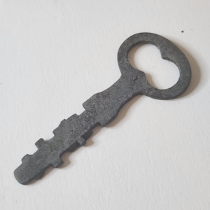 Könnte beinhalten: Ein dunkelgrauer, verwitterter Metallschlüssel mit einer texturierten, matten Oberfläche. Der Schlüsselkopf ist oval geformt mit einer zentralen Öffnung, und der Bart weist eine Reihe markanter, gezackter Zähne auf, die Altersspuren zeigen.