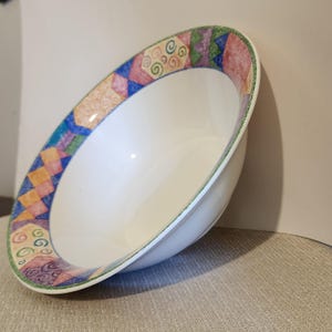 May include: A white ceramic bowl featuring a wide rim decorated with a colorful geometric pattern. The rim displays alternating shapes in shades of blue, orange, pink, green, and purple, accented with small spiral designs and a thin green border.