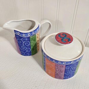 May include: A ceramic creamer pitcher and a lidded sugar bowl featuring a colorful vertical striped pattern in blue, green, orange, and purple. The pieces are decorated with white swirling line accents and a red circular lid knob with a blue design.