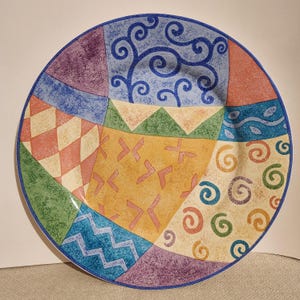 May include: A round ceramic plate featuring a vibrant patchwork design of various geometric shapes and patterns. The surface displays sections of blue swirls, orange diamonds, green triangles, yellow markings, and colorful spirals on a textured base.