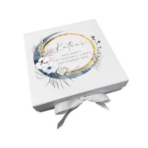 May include: White gift box with a gold and blue floral design. The box has a white satin ribbon and says "Katie's Hen Party Tattershall Lakes September 2022" on the lid.