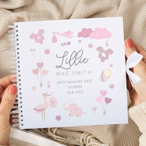 May include: A white baby memory book with a pink and white design. The book has a spiral binding and a white ribbon. The cover features a pink and white illustration of a baby, a stork, a cloud, a balloon, and a bottle. The book also includes the text "Lillie Mae Smith", "20th August 2022", "12:15PM", and "5LB 4OZ".
