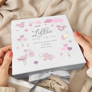 May include: A white gift box with a pink and white design featuring a stork, elephant, balloons, and other baby-themed illustrations. The box is tied with a silver ribbon and has the text "Lillie Mae Smith, 20th August 2022, 12:15PM, 5LB 4OZ" printed on it.