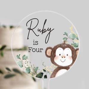 May include: A clear, round cake topper with the text "Ruby is Four" in black script. A cartoon monkey with brown fur and a smiling face is on the right. Green and gold foliage surrounds the edges. A tiered cake is in the background.