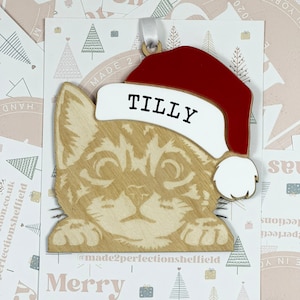 May include: A wooden cat ornament with a red Santa hat. The cat is wearing a white name tag that says "TILLY". The ornament is decorated with a "Merry Christmas" message and a small Christmas tree.