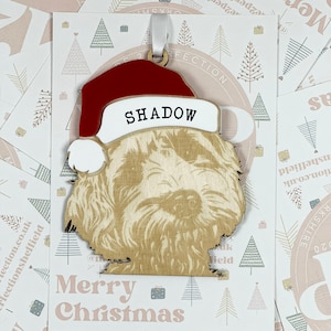 May include: A wooden Christmas ornament shaped like a dog wearing a Santa hat. The dog's name, "SHADOW", is written on the hat in white letters. The ornament has a white ribbon attached to the top.