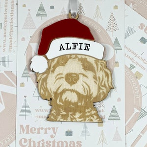 May include: A wooden Christmas ornament shaped like a dog wearing a Santa hat. The dog's name, "Alfie", is written on the hat. The ornament is on a white background with a pink and gray pattern.