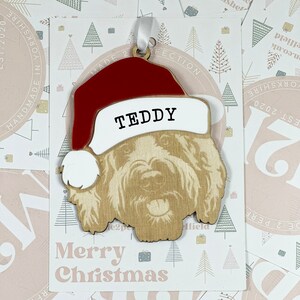May include: A wooden Christmas ornament shaped like a dog wearing a red Santa hat. The ornament has the name "TEDDY" written on the hat. The ornament is on a white background with a pink border and the words "Merry Christmas" printed on it.