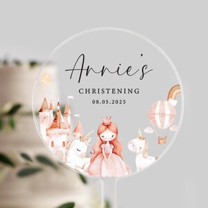 May include: A clear, circular cake topper with the text "Annie's Christening" and the date 08.05.2025. The design features a princess, unicorns, a castle, a hot air balloon, and a rainbow in soft pastel colours.