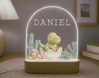 Puede incluir: Una luz nocturna de madera con un inserto de acrílico transparente que presenta un dinosaurio de dibujos animados en un nido con cactus y hojas. El nombre "DANIEL" está impreso en el inserto de acrílico.