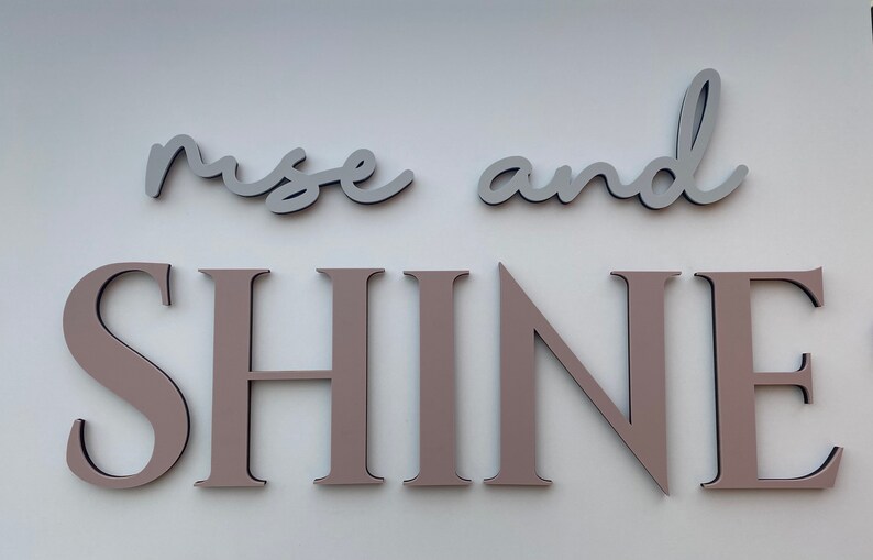 May include: Rise and Shine wall decor with the words "rise" and "and" in light gray and the word "SHINE" in a light brown color.