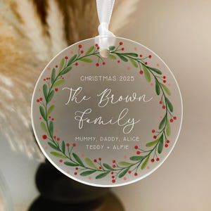 May include: A clear, round Christmas ornament with a white ribbon. The ornament features a green and red wreath design around the edge, with the text "CHRISTMAS 2025" and "The Brown Family" in elegant script. The names "Mummy, Daddy, Alice, Teddy + Alfie" are also included.