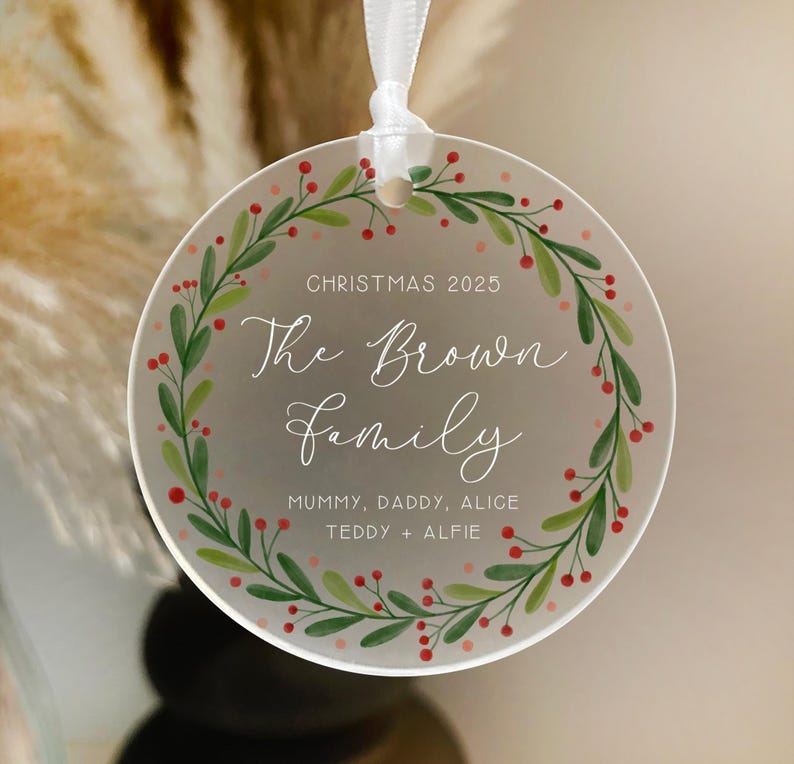 May include: A clear, round Christmas ornament with a white ribbon. The ornament features a green and red wreath design around the edge, with the text "CHRISTMAS 2025" and "The Brown Family" in elegant script. The names "Mummy, Daddy, Alice, Teddy + Alfie" are also included.