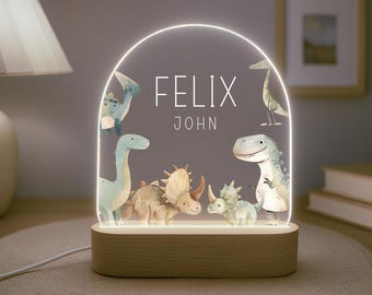Könnte beinhalten: Ein Nachtlicht aus Holz mit einer durchsichtigen Acrylscheibe mit Dinosaurier-Motiv. Die Scheibe hat einen weißen Rahmen und der Name "Felix John" ist in Weiß gedruckt. Das Licht ist eingeschaltet und zeigt verschiedene Dinosaurier im Aquarell-Stil.