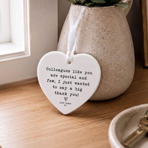 a heart-shaped ceramic tag hanging on a wooden surface, with a potted plant and keys visible in the background.