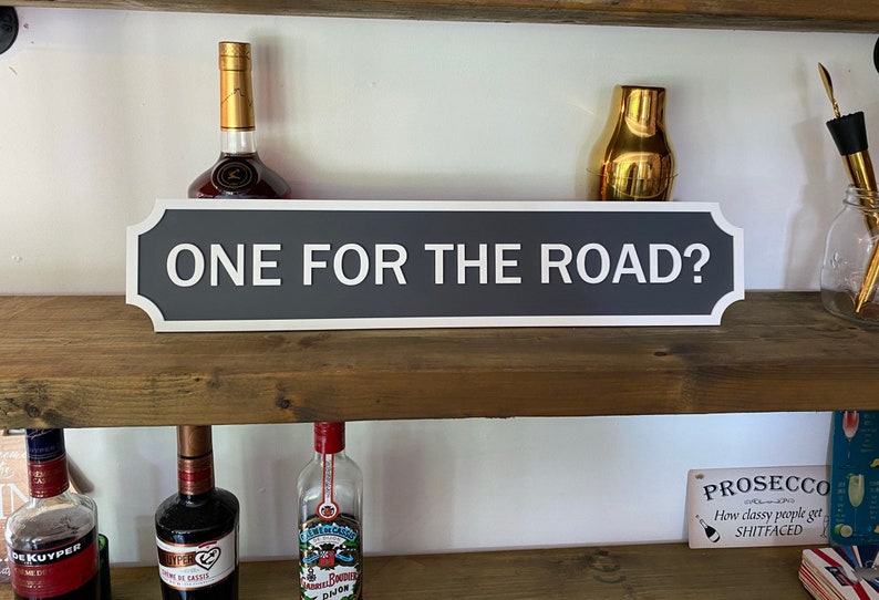 May include: A grey and white sign with rounded corners and a white border. The sign reads "ONE FOR THE ROAD?" and is displayed on a wooden shelf with various bottles and other items.