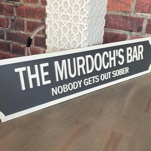 May include: A rectangular sign with the text "THE MURDOCH'S BAR" and "NOBODY GETS OUT SOBER" in white lettering. The sign has a gray center with a white border and is displayed against a brick wall.