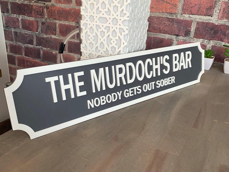 May include: A rectangular sign with the text "THE MURDOCH'S BAR" and "NOBODY GETS OUT SOBER" in white lettering. The sign has a gray center with a white border and is displayed against a brick wall.