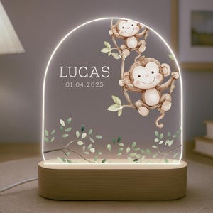 Może przedstawiać: Drewniana lampka nocna z przezroczystym panelem akrylowym przedstawiającym dwie kreskówkowe małpy huśtające się na winorośli. Panel jest podświetlany od tyłu, tworząc miękkie światło. Na panelu nadrukowany jest tekst "LUCAS 01.04.2025".