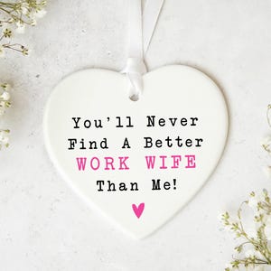 May include: A white heart-shaped ornament with the text "You'll Never Find A Better WORK WIFE Than Me!" in black and pink. A small pink heart is at the bottom. The ornament hangs from a white ribbon. White flowers are in the background.