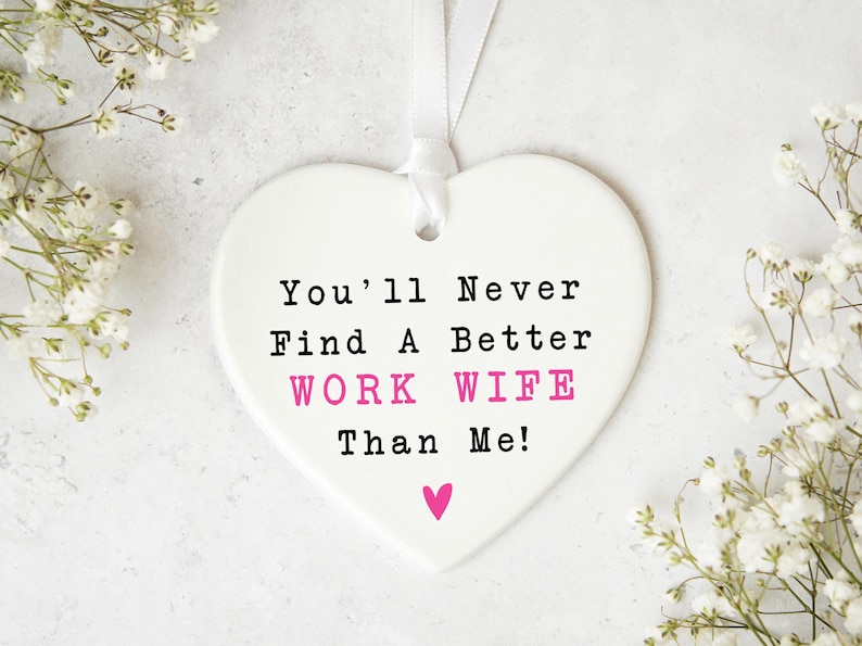 May include: A white heart-shaped ornament with the text "You'll Never Find A Better WORK WIFE Than Me!" in black and pink. A small pink heart is at the bottom. The ornament hangs from a white ribbon. White flowers are in the background.