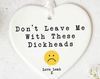 May include: A white heart-shaped ceramic ornament with the text "Don't Leave Me With These Dickheads" in black. A yellow sad face and the words "Love Leah X" are also on the ornament. A white ribbon is attached for hanging.