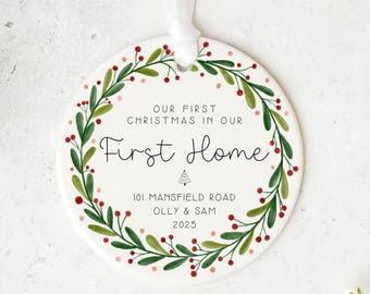 May include: A round, white ceramic Christmas ornament with a green and red wreath design. The ornament has the text "OUR FIRST CHRISTMAS IN OUR First Home" and the address "101 MANSFIELD ROAD OLLY & SAM 2025". A white ribbon is attached for hanging.