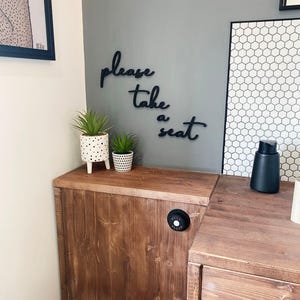 a bathroom with a wooden cabinet, a toilet, and a potted plant. There is a sign on the wall that says "Please take a seat".