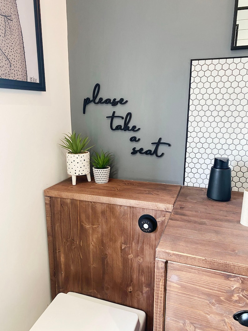 a bathroom with a wooden cabinet, a toilet, and a potted plant. There is a sign on the wall that says "Please take a seat".