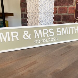 May include: A beige and white wooden sign with rounded edges and a white border. The sign reads "MR & MRS SMITH 02.08.2021".