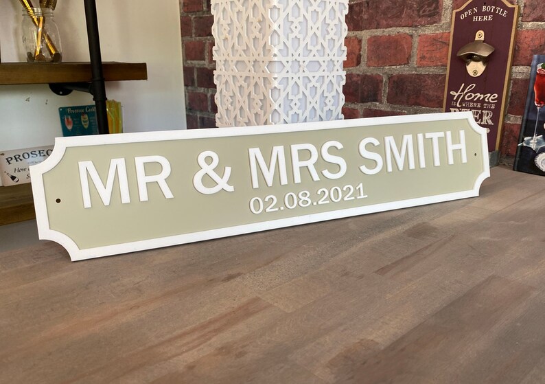 May include: A beige and white wooden sign with rounded edges and a white border. The sign reads "MR & MRS SMITH 02.08.2021".