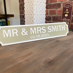 May include: A rectangular sign with the text "MR & MRS SMITH" and the date "02.08.2021" in white lettering. The sign has a light green background with a white border. The sign is displayed on a wooden surface.
