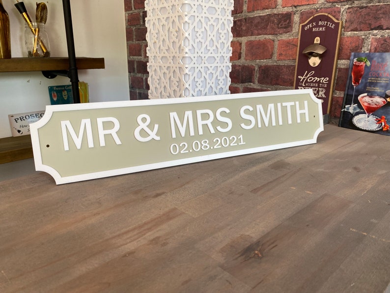 May include: A rectangular sign with the text "MR & MRS SMITH" and the date "02.08.2021" in white lettering. The sign has a light green background with a white border. The sign is displayed on a wooden surface.