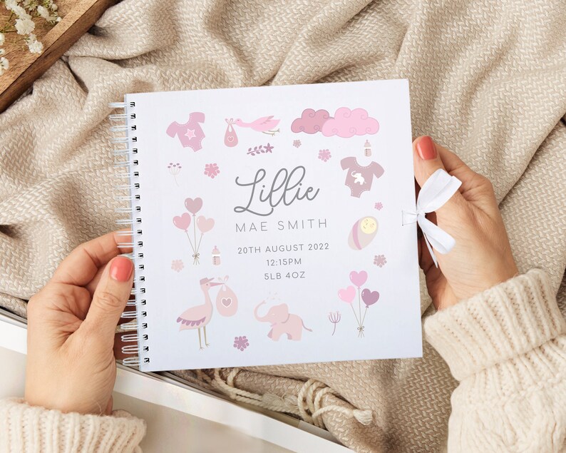 May include: A white baby memory book with a pink and white design. The book has a spiral binding and a white ribbon. The cover features a pink and white illustration of a baby, a stork, a cloud, a balloon, and a bottle. The book also includes the text "Lillie Mae Smith", "20th August 2022", "12:15PM", and "5LB 4OZ".