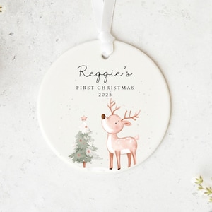 May include: A circular Christmas ornament with a ribbon, featuring a watercolour illustration of a reindeer and a Christmas tree. The text on the ornament reads "Reggie's First Christmas 2025".