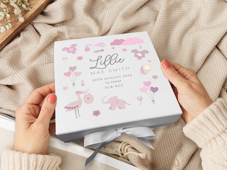 May include: A white gift box with a pink and white design featuring a stork, elephant, balloons, and other baby-themed illustrations. The box is tied with a silver ribbon and has the text "Lillie Mae Smith, 20th August 2022, 12:15PM, 5LB 4OZ" printed on it.