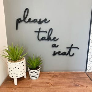 a wooden surface with two potted plants, and there is a sign on the wall that says "Please take a seat".