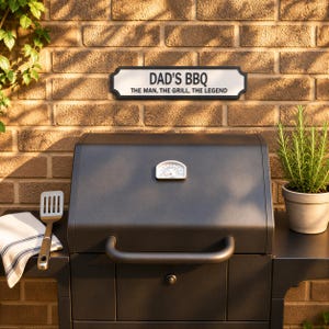 a black barbecue grill with a sign above it that reads "DAD'S BBQ THE MAN, THE GRILL, THE LEGEND". The grill is set against a brick wall, and there is a potted plant and a spatula nearby.