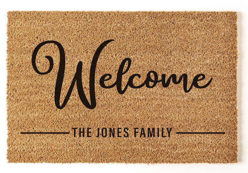 May include: A brown coir doormat with the text "Welcome" in a black cursive font. Below the text is a line with the text "The Jones Family" in black.